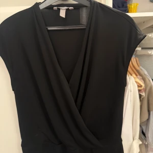 Svart omlottopp från H&M - Säljer en svart omlottopp från H&M i storlek S. Toppen har en djup v-ringning och är ärmlös med en snygg draperad omlottdesign framtill. Perfekt att styla med jeans eller kjol för en clean look.
