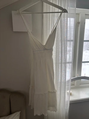 Vit somrig kortklänning med volang - Superfin vit kortklänning med smala axelband och v-ringning. Klänningen har volangdetaljer nertill och är tillverkad i ett lätt, luftigt material som passar perfekt för varma dagar. Snygg och enkel att styla med sandaler eller sneakers.