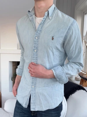 Ljusblå skjorta från Ralph Lauren - Säljer en ljusblå skjorta från Ralph Lauren i slim fit-modell. Skjortan har klassisk krage, knappar framtill och det ikoniska broderade logotypen på bröstet. Tillverkad i mjuk bomull och har långa ärmar med knapp vid manschetten. Perfekt till jeans eller chinos.