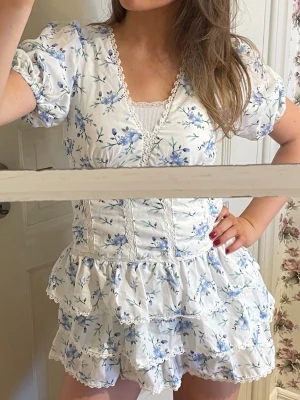 Blommig vit kortklänning med volanger - Köpte på Sellpy, men tyvärr var den för liten och kort för mig som är 166cm. Supersöt kortklänning i vitt med blått blommönster och puffärmar. Klänningen har v-ringning med spetsdetaljer, markerad midja och flera lager volanger i kjolen. Lappar kvar!