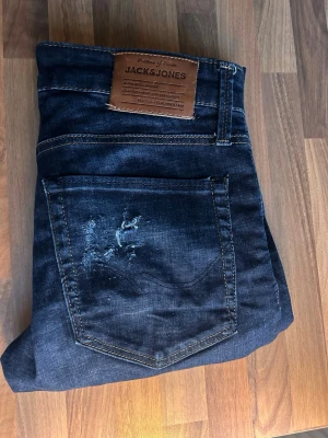 Mörkblå Slim Glenn jeans Jack & Jones - Mörkblå jeans från Jack & Jones, modell Slim/Glenn. Jeansen har klassisk femficksdesign, slitningar på framsidan och kontrastsömmar. Tillverkade i stretchig bomullsdenim för skön passform och en modern slim siluett.