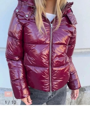 Glansig vinröd pufferjacka - Säljer en glansig vinröd pufferjacka från Nelly med huva och dragkedja framtill. 
