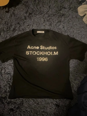 Svart Acne Studios t-shirt Stockholm - Svart t-shirt från Acne Studios med tryck 'Acne Studios STOCKHOLM 1996' på bröstet. Klassisk passform och rund hals. Tillverkad i 100% bomull för en skön känsla. Perfekt för dig som gillar stilrena och ikoniska plagg. Storlek s. Lappen finns använd några få gånger