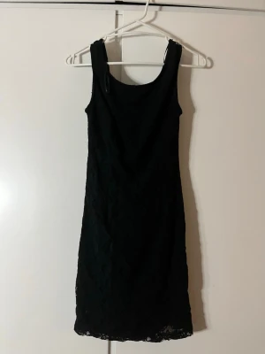 Svart spetsklänning med ärmlös design - Säljer en svart kortklänning med spetsdetaljer och ärmlös design. Klänningen har en rundad halsringning fram och djupare ringning i ryggen. Perfekt för dig som gillar enkel men elegant stil.