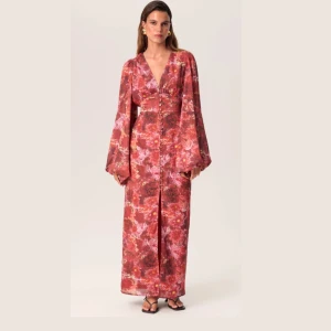 Blommig långklänning från Adoore - Populär långklänning från Adoore i varma rödbruna toner med blommigt mönster. Klänningen har v-ringning, långa puffärmar och dekorativa knappar framtill. 