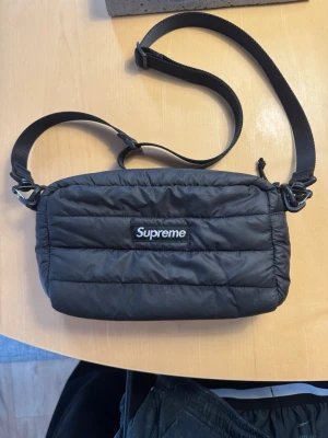 Supreme svart quiltad axelväska - Supreme axelväska i svart med quiltad design och justerbar rem. Väskan är tillverkad i lätt och slitstark Pertex Quantum polyester och har dragkedja upptill samt meshficka inuti. Perfekt för att bära dina essentials med stil. Väldigt ovanlig! 