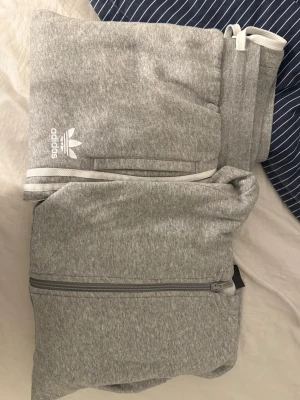 Adidas set - Ett jättefint adidas set i grå och vit, storlek 170 i byxor och 164 i tröja. Tröjan är en huvtröja med text på ärmen och även byxorna har märke och adidas ränder. Går att köpa både enskilt och i set! Bra skick och inte synligt använd. Hör av dig om du vill köpa enskilt!