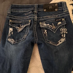 Miss Me jeans med paljettdetaljer - Lowwaist mörkblå jeans från Miss Me med bootcut-modell  och paljettdetaljer där bak. Tydliga kontrastsömmar och broderad logga. De enda defekterna som finns är längst ner på benen som jag tagit bild på men det är inget som syns när man har på sig dem. Eftersom de är vintage säljer jag nära (nypris 1499kr) trots defekterna, har kvittot om ni vill se. De är köpta på DEPART A MENTAL A2 vintage och använda få gånger. Skriv privat för fler bilder!⭐️ tyvärr lite för små för mig annars hade jag behållt!