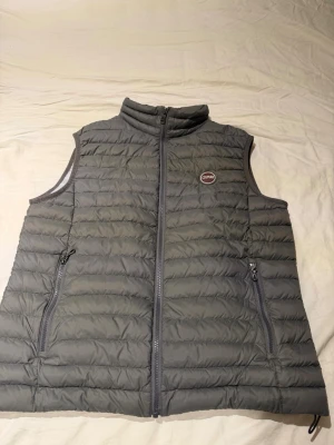 Grå dunväst från Colmar - Säljer en stilren grå dunväst från Colmar med quiltad design och hög krage. Västen har två dragkedjeförsedda fickor och en tydlig Colmar-logga på bröstet. Perfekt lager för kyliga dagar och enkel att matcha med olika outfits.