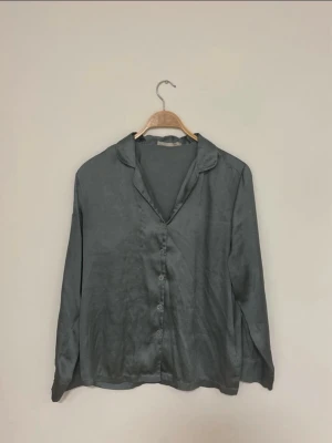 Mörkgrå sidenväst från 157 - Säljer en mörkgrå tunna tröja från 157 i mjukt sidenmaterial. Tröjan har klassisk krage, knäppning framtill med flera knappar och en enkel, stilren look. Perfekt att gå hemma o mysa med framför filmen.