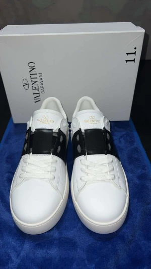 Valentino Garavani Open sneakers - Snygga Valentino Garavani Open sneakers i vitt skinn med bred svart läderdetalj över mitten och vita snören. Rund tå och platt sula med mönstrad undersida. Ikonisk logga på plösen och boxen medföljer. Perfekt för dig som gillar stilrena designer.