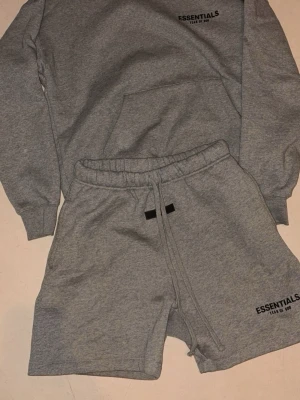 Grå Essentials shorts med snörning - Säljer ett par grå mjukisshorts från Essentials Fear of God. De har elastisk midja med långa snören och logga tryckt på ena benet. Perfekta för chill dagar och har en loose fit som ger skön streetkänsla.