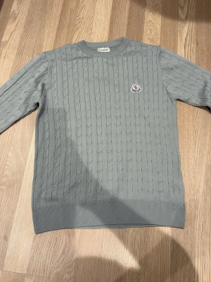 Grå kabelstickad tröja från Moncler - Säljer en grå kabelstickad tröja från Moncler med rund hals och diskret logga på bröstet. Tröjan har lång ärm och ribbade muddar vid ärmslut och nederkant. Perfekt för dig som gillar stilrena och klassiska plagg.