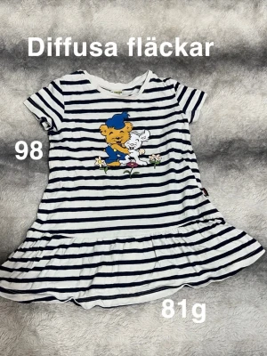 Randig tunika med Bamse-tryck - Söt tunika med ränder samt ett färgglatt Bamse-tryck framtill. Kortärmad modell med rund hals och volangkant nedtill. Perfekt för dig som gillar lekfulla motiv och mjuka material.🌸 Diffusa fläckar. Storlek 98