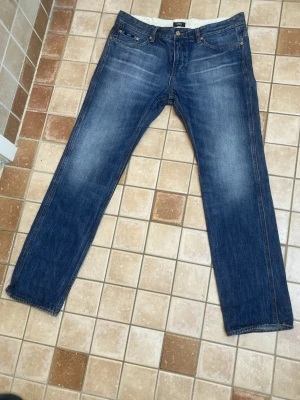 Blå jeans från Hugo Boss Ice Wash - Säljer ett par blå jeans från Hugo Boss med Ice Wash-effekt som ger en fräsch och mjuk indigo-färg. Klassisk femficksmodell med raka ben och kontrastsömmar på bakfickan. Materialet är slitstarkt jeans och passformen är normal. Knappt använda. Storlek 34/32.