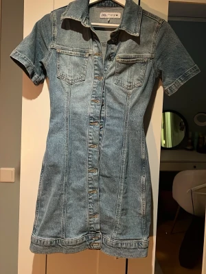Kort jeansklänning från Zara - Säljer en snygg kort jeansklänning från Zara med korta ärmar, krage och knappar hela vägen framtill. Klänningen har två bröstfickor och markerade sömmar som ger en cool vintagekänsla. Perfekt för dig som gillar denim och vill ha en avslappnad men trendig look.