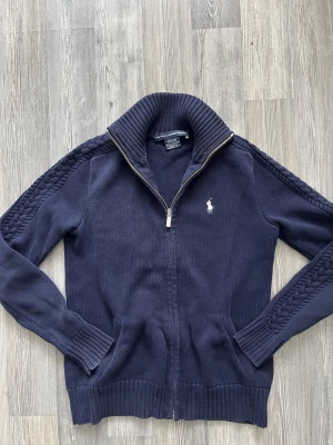 Mörkblå stickad kofta från Ralph Lauren - Snygg mörkblå stickad kofta från Ralph Lauren med dragkedja och ribbad krage. Tröjan har kabelstickade detaljer längs ärmarna och klassisk logga broderad på bröstet. Tillverkad i 100% bomull och har långa ärmar med ribbade muddar.