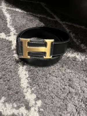 H-bälte  - Snyggt bälte i guld och svart skinn med ikoniskt,H-spänne i guldfärgad metall. Bältet har stilrena, kontrastsömmar och klassisk design som lyfter, vilken outfit som helst. Strl 110cm