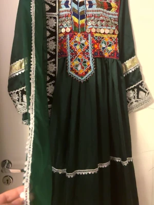 Afghansk klänning med byxa och sjal - Unik långklänning i mörkgrönt med färgstarka broderier och silverfärgade paljettdetaljer. Klänningen har långa ärmar med broderade mönster och spetsdetaljer, samt matchande byxa och sjal med paljettkantPerfekt för dig som vill sticka ut med traditionell och färgglad stil.