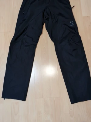 Svarta täckbyxor från Haglöfs - Säljer ett par svarta täckbyxor från Haglöfs med elastisk midja och justerbar dragsko. Byxorna har dragkedjeförsedda fickor och är tillverkade i ett slitstarkt syntetmaterial, perfekta för utomhusaktiviteter. Snygg och funktionell design med diskret logga på benet.