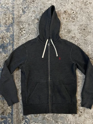 Mörkgrå hoodie från Polo Ralph Lauren - Mörkgrå hoodie från Polo Ralph Lauren med dragkedja, vita snören och klassisk röd logga på bröstet. Mjuk insida och känguruficka framtill. Perfekt för chill dagar eller när du vill ha en clean streetstyle.