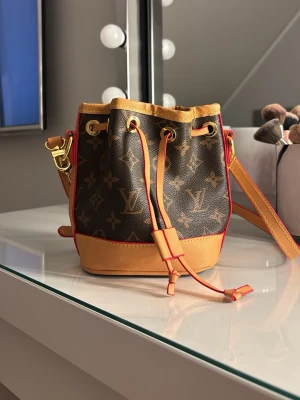 Bucket bag  - Säljer en snygg axelväska  i klassiskt monogrammönster. Väskan är brun med ljusbruna och röda detaljer, har justerbar axelrem och stängs med snörning. Materialet är skinn och canvas, perfekt för dig som gillar lyxiga accessoarer.