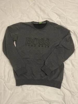 Grå sweatshirt från Hugo Boss - Säljer en stilren grå sweatshirt från Hugo Boss med svart logga på bröstet. Tröjan har rund halsringning, ribbade muddar och är tillverkad i mjuk bomullsblandning. Perfekt till jeans eller joggers! Denna kan även passa S! Har inga defekter.