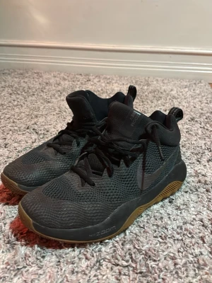 Svarta Nike Zoom Rev sneakers - Säljer ett par svarta Nike Zoom Rev sneakers med högt skaft och bruna gummisulor. Skorna har mesh-överdel för bra ventilation, snörning och en stor Nike-logga på sidan och undersidan. Perfekta för dig som gillar sportig stil och vill ha bekväma skor.