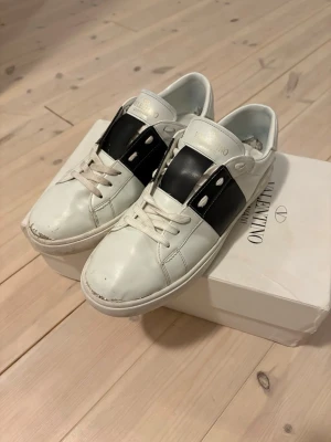 Valentino Garavani Open sneakers vit/svart - Säljer ett par Valentino Garavani Open sneakers i vitt skinn med bred svart läderdetalj över mitten. Klassisk rund tå, vita snören och diskret logga på plösen. Snygg och stilren design som passar till det mesta. Kommer med originalkartong och påse. Vid snabb affär så kan pris gå ner mycket