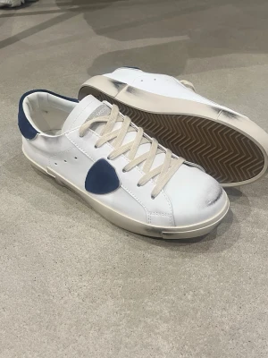 Vita sneakers från PHILIPPE MODEL - Snygga vita sneakers från PHILIPPE MODEL med marinblå detaljer på sidan och hälen. Skorna har beige snörning, bruna yttersulor och coola slitningar på sulan för en trendig look. Tillverkade i skinn med klassisk låg profil och rund tå.