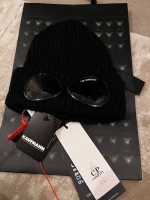 Svart mössa med goggles från C.P. Company - Ribbstickad svart mössa från C.P. Company med ikoniska inbyggda goggles framtill. Mössan är tillverkad i mjukt material och har en uppvikt kant. Perfekt accessoar för att sticka ut och hålla värmen under kalla dagar.