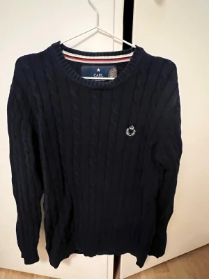 Mörkblå kabelstickad tröja  - Säljer en mörkblå kabelstickad tröja från Carl med rund halsringning och diskret broderad logga på bröstet. Tröjan har långa ärmar och ribbade muddar vid ärmslut och nederkant. Perfekt för kyliga dagar!