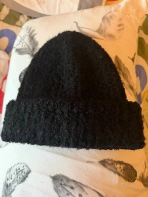 Svart boucle mössa från Stockh LM Studio - Säljer en svart boucle mössa från Stockh LM Studio. Mössan har en fluffig och texturerad yta med uppvikt kant, vilket ger en cool och trendig look. Perfekt för dig som gillar att sticka ut med accessoarer. Passar till både street och chill.