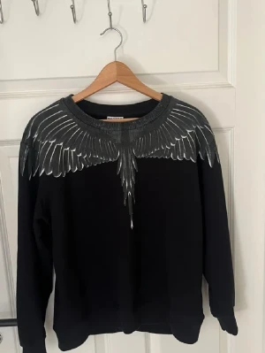 Svart tröja Marcelo Burlon med vingar - Svart sweatshirt från Marcelo Burlon County of Milan med ikoniskt vingmönster i grått och vitt över axlar och bröst. Tröjan har rund halsringning och långa ärmar. Perfekt statement-plagg för dig som gillar streetwear och unika prints.