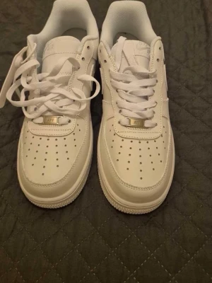 Nike Air Force 1 helvita sneakers - Klassiska Nike Air Force 1 i helvitt skinn med perforerad tå, vit sula och vita skosnören. Snygg låg modell med metallbricka på snörningen där det står 'AF1'. Perfekta till streetwear och passar året runt.