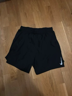 Svarta Nike shorts  - Säljer ett par svarta Nike shorts med elastisk midja och snörning. Klassisk loose fit och diskret Nike-logga i vitt på benet. Perfekta för träning eller chill dagar. Lätta och bekväma, tillverkade i syntetmaterial.
