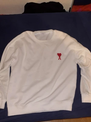 Vit sweatshirt från Ami Paris med rött hjärta - Säljer en clean vit sweatshirt från Ami Paris med det ikoniska röda hjärta-A broderiet på bröstet. Tröjan har rund hals, långa ärmar och är gjord i mjuk bomull. Perfekt för dig som gillar stilrena och trendiga plagg.