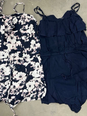 Två jumpsuits med smala axelband - Säljer två snygga jumpsuits med smala axelband. Ena är marinblå med volang upptill och snörning i midjan, den andra har ett blommigt mönster i vitt, rosa och mörkblått med snörning i midjan. Perfekt för sommarens häng och chill.