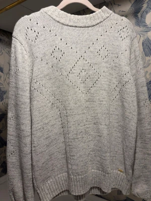 Ljusgrå stickad tröja med hålmönster - Superskön ljusgrå stickad tröja med snyggt hålmönster i diamantform på framsidan. Tröjan har ribbade muddar vid ärmslut och nederkant samt rundad hals. Perfekt för kyliga dagar och enkel att styla med jeans eller kjol.