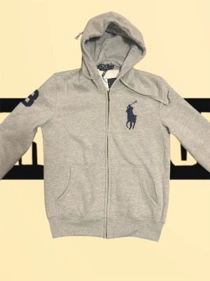 Grå Ralph lauren hoodie  - Stilren och grisch hoodie från Polo Ralph Lauren i ljusgrå färg, storlek M. Tröjan är i mycket bra skick utan synliga skador eller fläckar. Tillverkad i mjuk bomullsblandning, perfekt för både vardag och avslappnade tillfällen. Utrustad med dragkedja, fickor framtill och klassisk huva samt det ikoniska Polo-logot på bröstet och siffra på ärmen. Passformen är normal och bekväm.