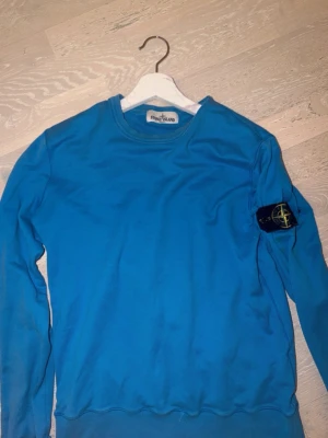 Blå sweatshirt från Stone Island Äkta Storlek M - Säljer en blå sweatshirt från Stone Island med den klassiska patchen på ärmen. Tröjan har rund halsringning och är tillverkad i mjuk bomull. Perfekt för dig som vill ha en clean och stilren look med streetwear-vibe.