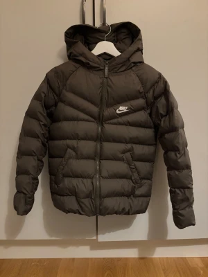 Snygg barnjacka från Nike - Säljer en grön  pufferjacka från Nike med huva och dragkedja framtill. Jackan har två fickor med knappstängning och en klassisk Nike-logga på bröstet. Perfekt för kalla dagar och riktigt skön att bära. Passar dig som gillar sportig och clean stil.