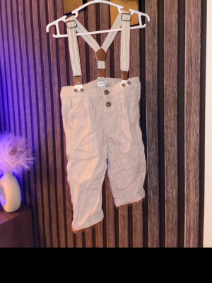 Newbie Beige byxor med hängslen storlek 68 - Säljer ett par newbie beige byxor med snygga justerbara hängslen och knappar framtill. Byxorna har uppvikta benslut och är gjorda i ett mjukt bomullstyg. Perfekt för en chill och stilren look. Storlek 68