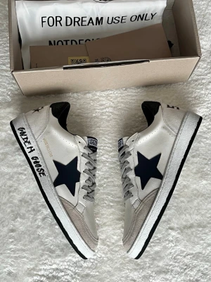 Golden Goose vita sneakers med svart stjärna - Golden Goose sneakers i vitt skinn med svart stjärna på sidan och svart häl. Grå snörning, vit sula med svart text och detaljer i beige mocka framtill. Klassisk låg modell med rund tå och perforeringar på tån för extra stil.