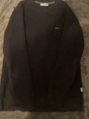 Svart långärmad tröja från Lacoste - Svart långärmad tröja från Lacoste i 100% bomull. Klassisk rund halsringning och diskret Lacoste-logga på bröstet. Perfekt basic-plagg med clean look och skön passform.