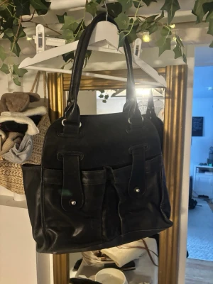 Svart skinnhandväska från Argentina - Snygg svart handväska i kanske äkta skinn från Argentina. Väskan har två stora ytterfickor med tryckknappar och långa handtag. Insidan har dragkedjefack för småsaker. Perfekt för dig som vill ha en stilren och rymlig väska med klassisk look.