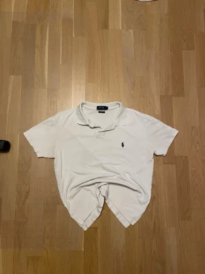 Vit piké från Polo Ralph Lauren - Klassisk vit pikétröja från Polo Ralph Lauren i storlek L men custom fit så passar 170-180. Tröjan har korta ärmar, krage och två knappar vid halsen. Ikoniska mörkblå loggan broderad på bröstet. Tillverkad i mjuk bomull som andas.