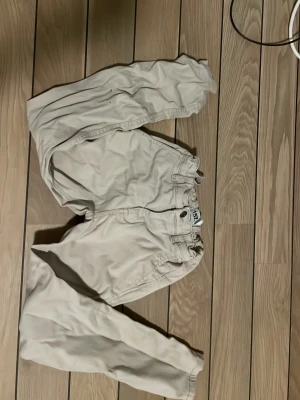 Beiga chinos från 157 - Säljer ett par klassiska beiga chinos från 157. Byxorna har normal passform, raka ben och midja med knapp och dragkedja. Tillverkade i mjuk bomull som är sköna att bära hela dagen. Perfekta till en avslappnad stil.