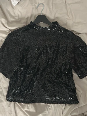 Svart paljettopp med kort ärm - Säljer en svart topp täckt med glittriga paljetter. Modellen har korta ärmar och en högre halsringning. Perfekt för dig som vill sticka ut och glänsa lite extra. Tyget är mjukt och följsamt, vilket gör den bekväm att bära hela kvällen.