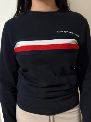 Svart sweatshirt från Tommy Hilfiger - Säljer en svart sweatshirt från Tommy Hilfiger med klassiska ränder i marinblått, vitt och rött över bröstet. Tröjan har rund halsringning och långa ärmar. Perfekt för en avslappnad och stilren look.